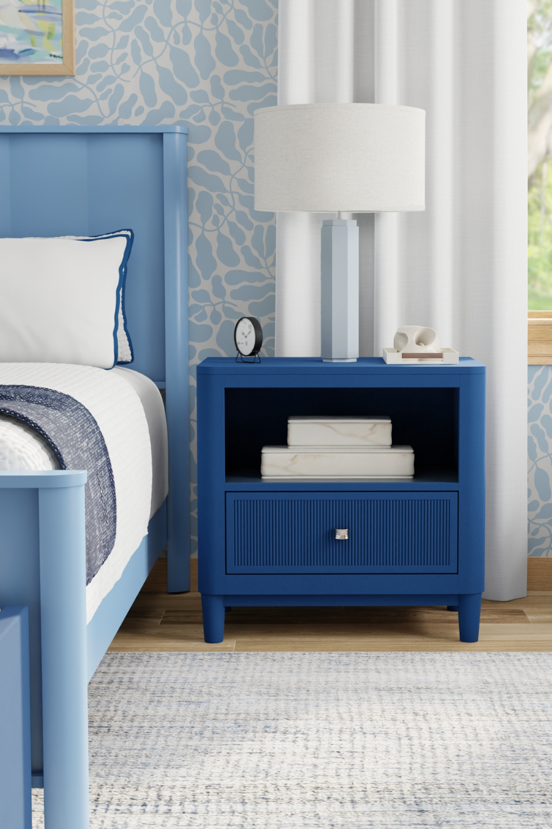 Blue nightstand with books and a lamp next to a bed with blue headboard and white bedding.